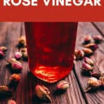 A glass jar of rose vinegar on a wooden table with dried rosebuds scattered around; text reads “How to Make Rose Vinegar.”.