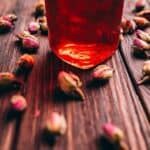 A close-up of a glass jar with red liquid on a wooden surface, surrounded by dried rosebuds.