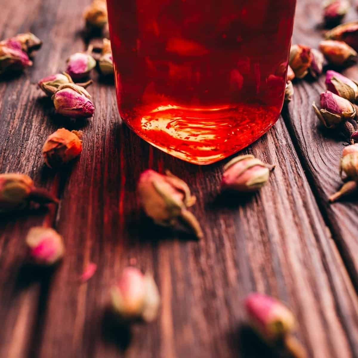 A close-up of a glass jar with red liquid on a wooden surface, surrounded by dried rosebuds.