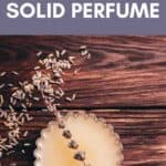 A small tin of solid perfume infused with lavender sits on a wooden surface, with dried lavender and seeds scattered around. Text above reads, “DIY Lavender Solid Perfume.”.