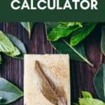 A bar of soap topped with a leaf sits on a wooden surface surrounded by green leaves. Text reads “Melt and Pour Soap Essential Oil Calculator,” highlighting the perfect tool for anyone seeking an easy essential oil calculator to craft their own custom-scented soaps.