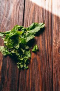A small pile of fresh spinach leaves sits on a dark wooden surface, bathed in natural light—perfect alongside a cup of fresh mint tea.