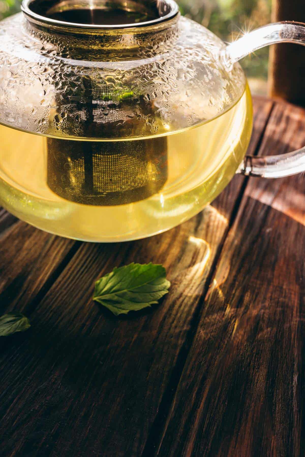A glass teapot with yellow-green fresh mint tea sits on a wooden table, with a few green leaves placed nearby.
