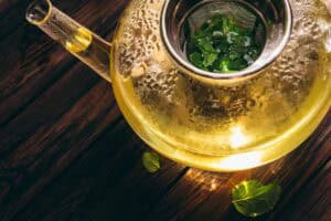Glass teapot with fresh mint tea steeping in hot water on a wooden table, viewed from above.