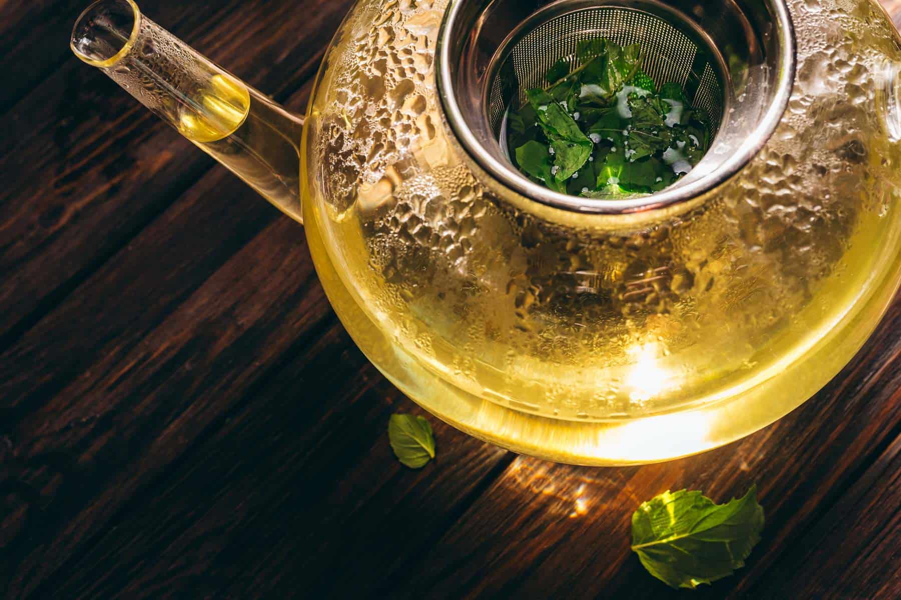 Glass teapot with a metal infuser brewing fresh mint tea on a wooden table. Light reflects off the teapot, and a few mint leaves are scattered nearby.