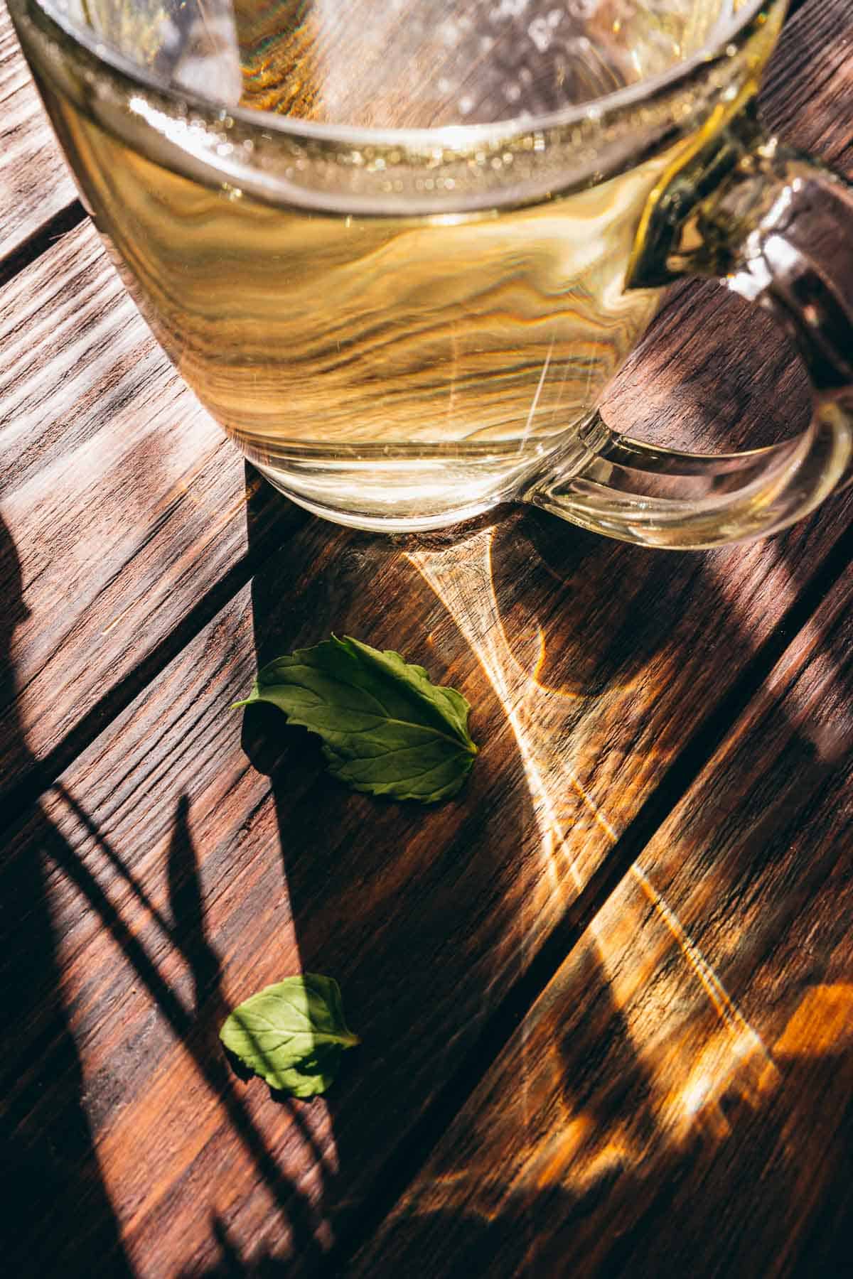 Glass cup filled with fresh mint tea on a wooden table with two mint leaves beside it. Sunlight casts shadows and reflections across the scene.