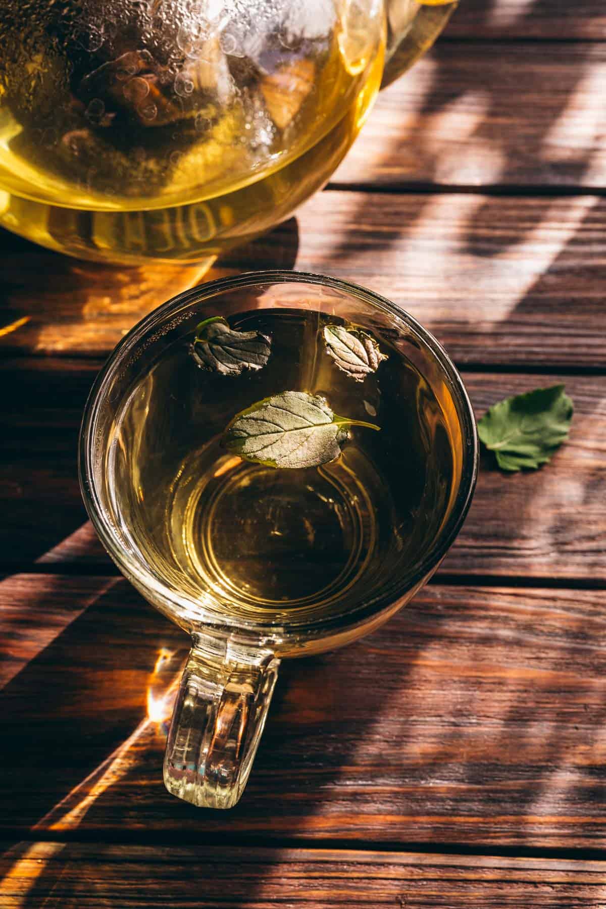 A clear glass cup of fresh mint tea with mint leaves floating on the surface, placed on a wooden table with sunlight streaming in.