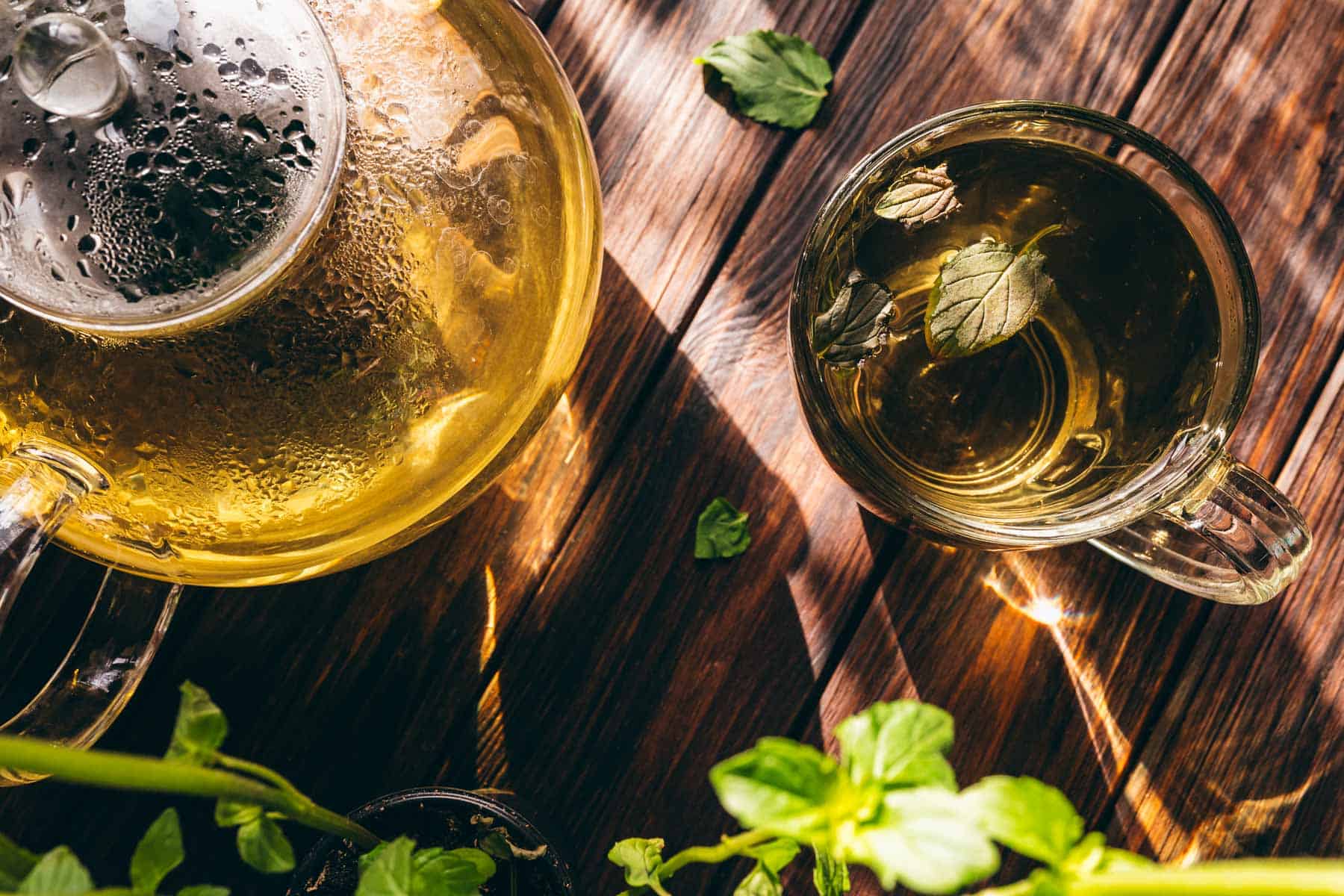 Glass teapot and teacup with hot fresh mint tea on a wooden table, scattered mint leaves, and sunlight casting gentle shadows.