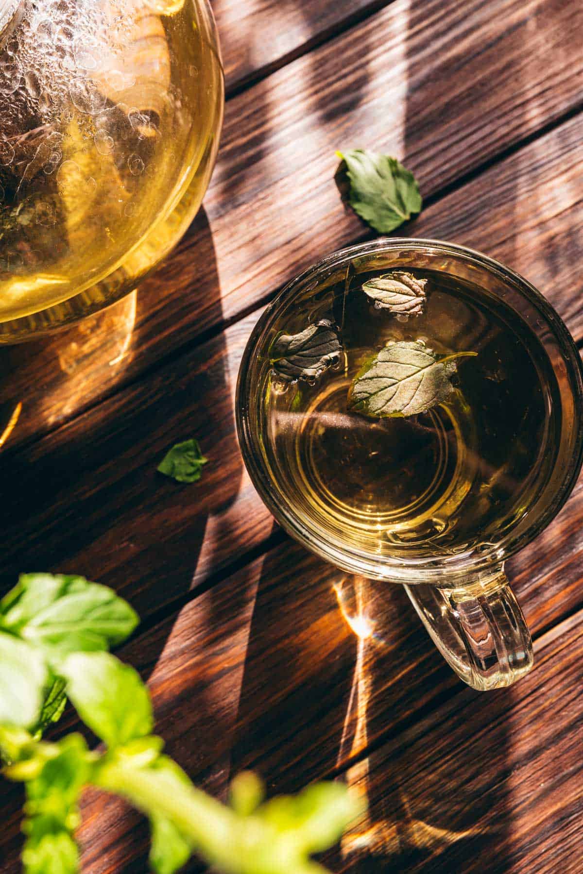 A glass cup of fresh mint tea with mint leaves sits on a wooden table, next to a glass teapot and scattered mint leaves, in natural sunlight.
