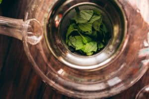 Top view of fresh mint tea leaves inside a strainer placed in a glass teapot on a wooden surface.