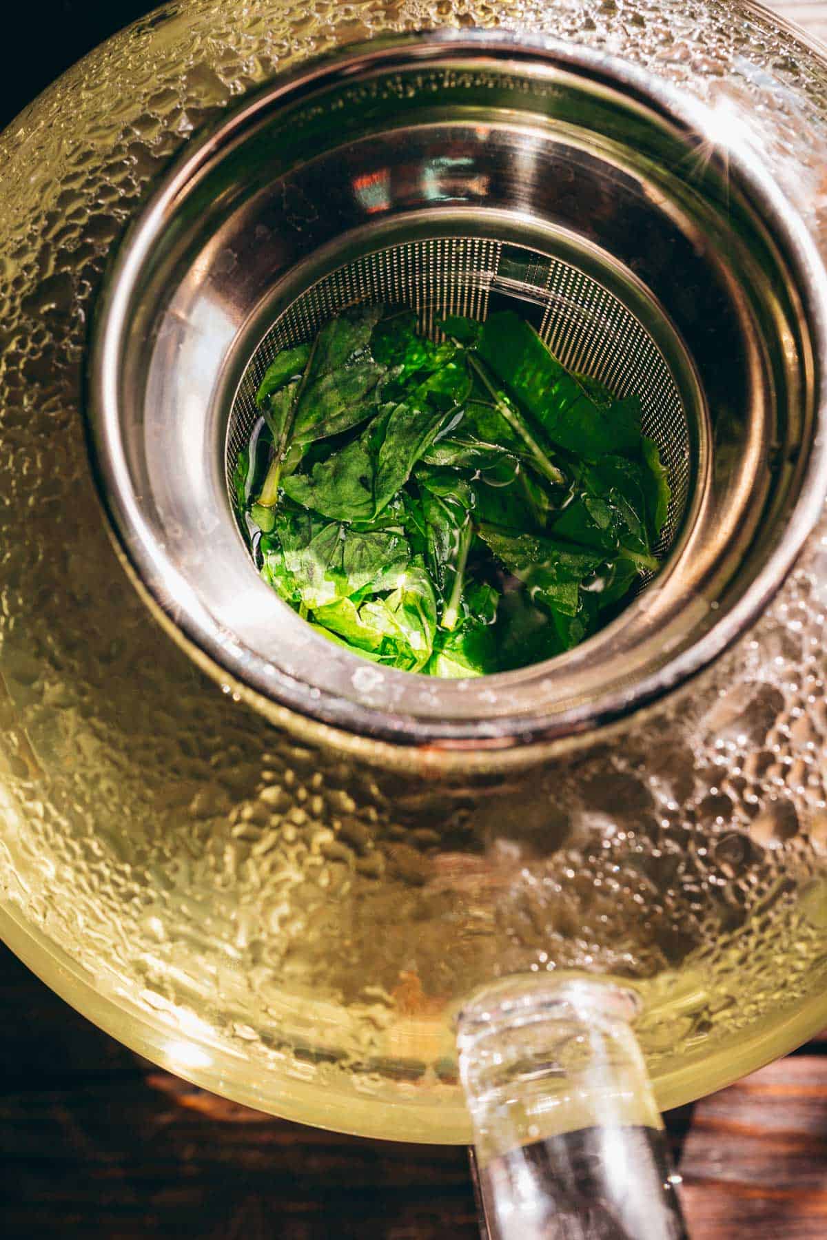 Top view of fresh mint tea leaves inside a metal infuser in a glass teapot, with steam droplets visible on the teapot’s surface.
