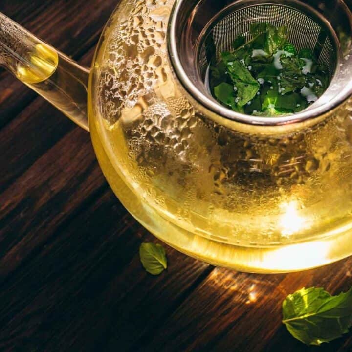 Glass teapot with strainer holding fresh mint tea leaves, filled with hot water, sits on a wooden surface with a few loose mint leaves nearby.