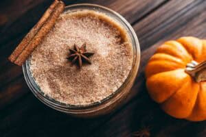 A glass jar filled with brown sugar scrub, topped with a cinnamon stick and star anise, sitting next to a pumpkin.