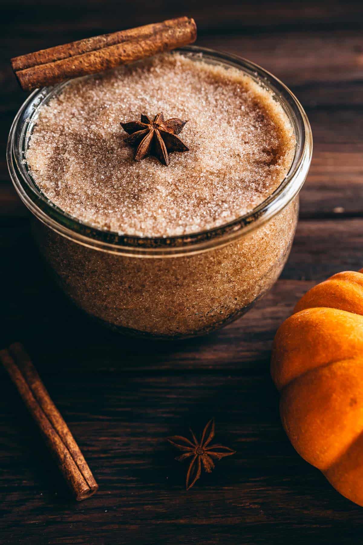 Glass jar filled with pumpkin spice sugar scrub, topped with a star anise, alongside a cinnamon stick and a pumpkin.