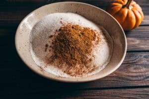 A bowl of white sugar topped with ground cinnamon, next to a small pumpkin on a wooden surface.