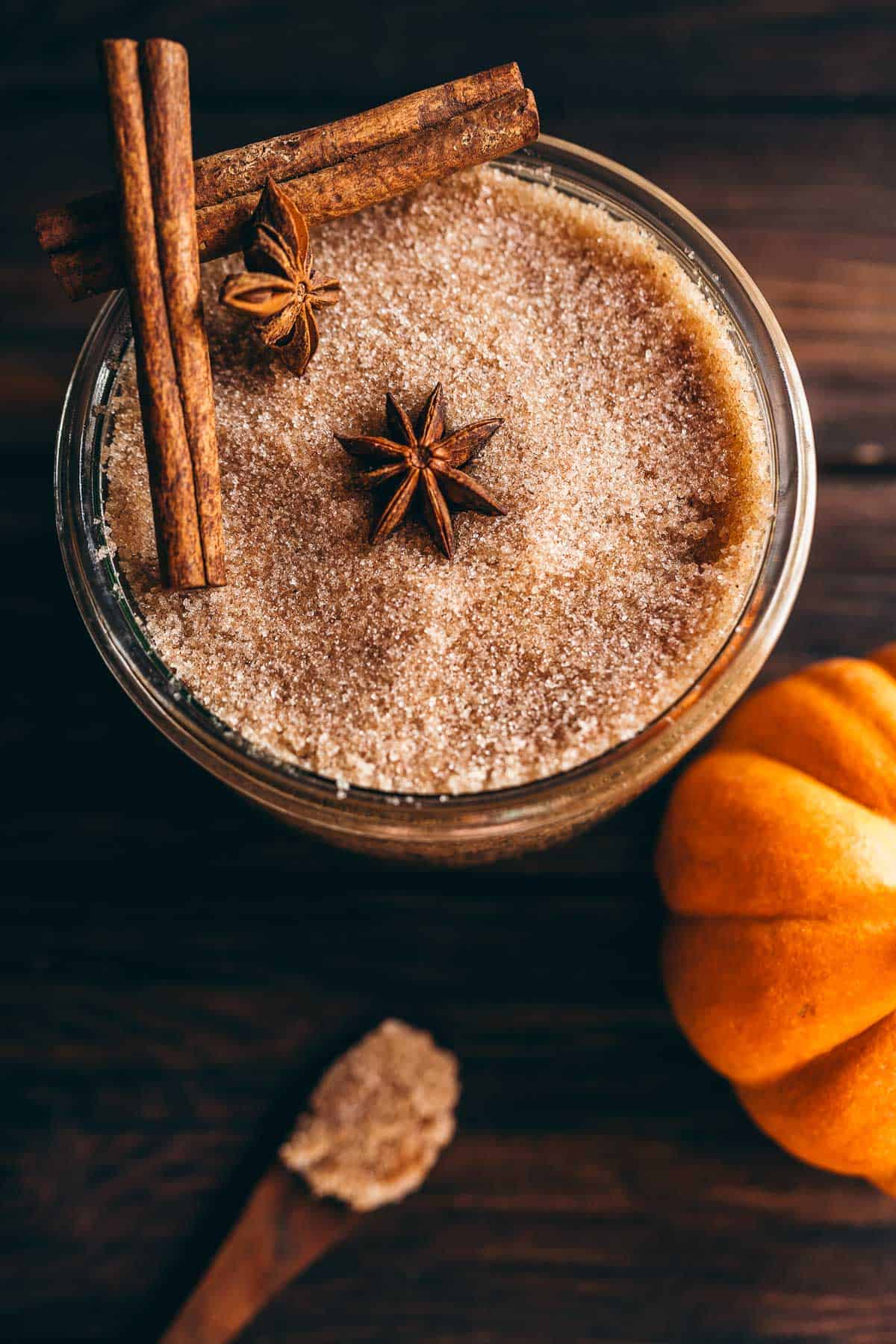 A glass jar filled with pumpkin spice sugar scrub, topped with cinnamon sticks and star anise, sitting next to a small pumpkin.