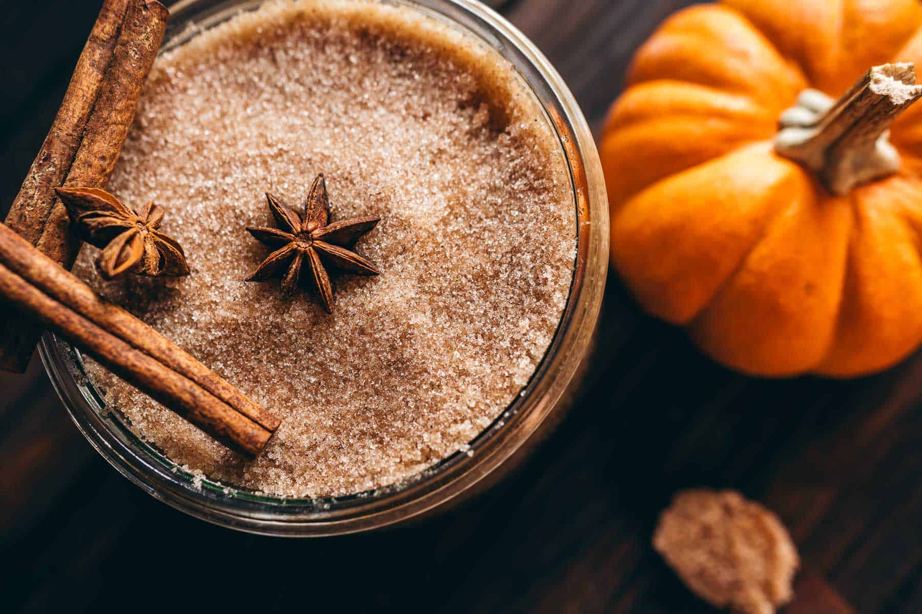 A glass jar filled with pumpkin spice sugar scrub, topped with cinnamon sticks and star anise, next to a small pumpkin.