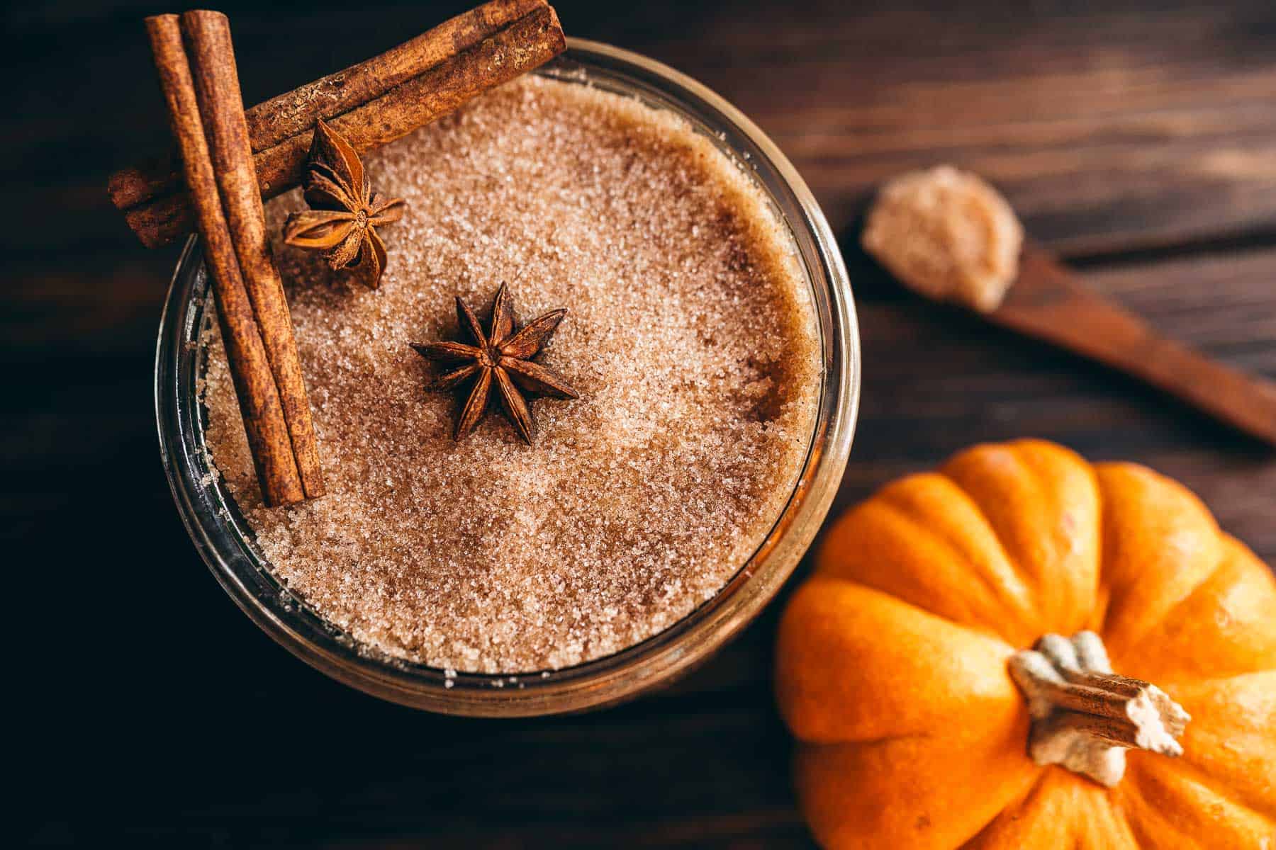 A jar of pumpkin spice sugar scrub topped with cinnamon sticks and star anise, next to a small pumpkin.