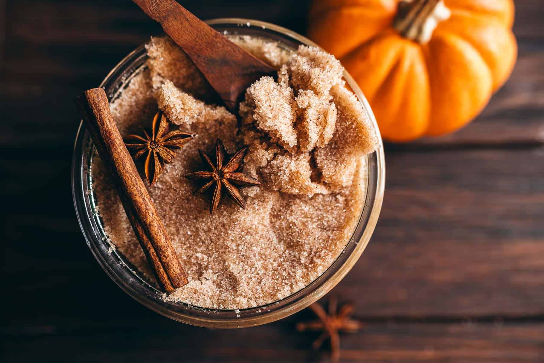 A glass jar of pumpkin spice sugar scrub topped with a wooden spoon, a cinnamon stick, and two star anise, set beside a small pumpkin.