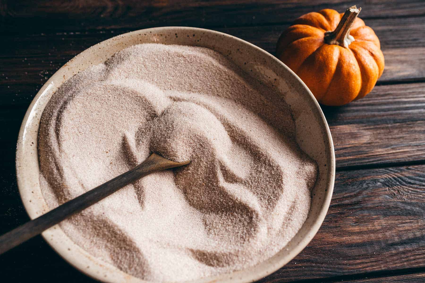A bowl of pumpkin spice sugar scrub with a wooden spoon, next to a small pumpkin on a dark wooden surface.