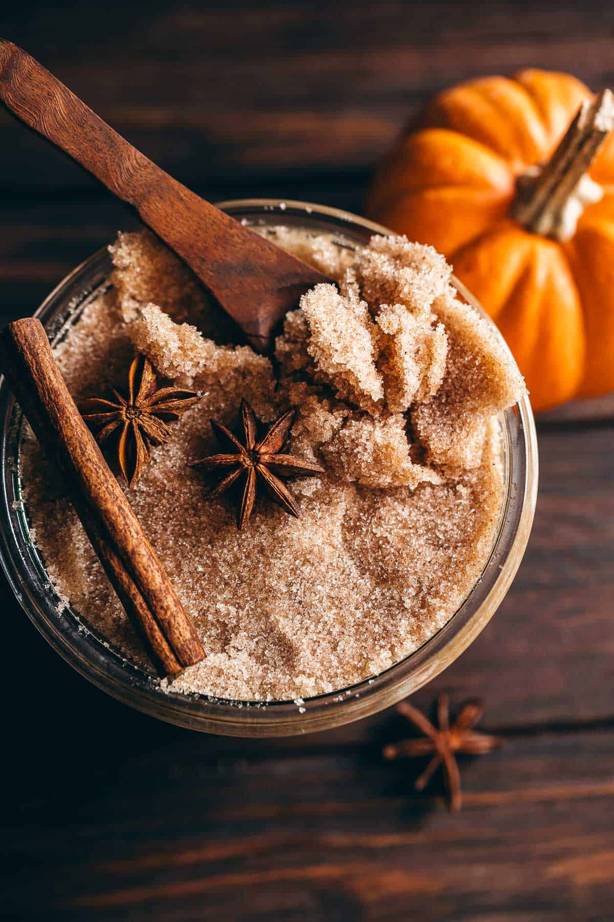 A jar of pumpkin spice sugar scrub topped with two star anise and a cinnamon stick, with a wooden spoon and a small pumpkin nearby.