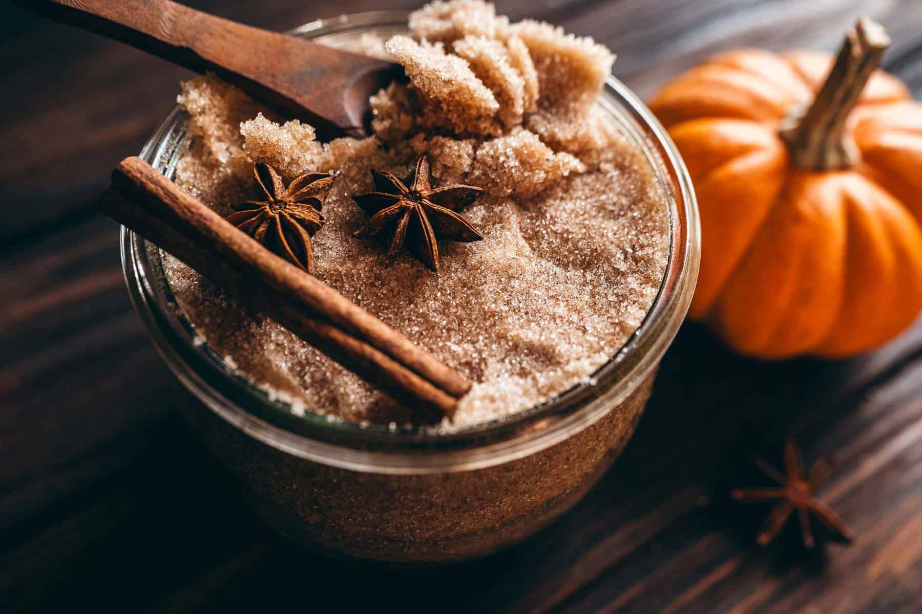 Glass jar filled with pumpkin spice sugar scrub, topped with a wooden spoon, two star anise, and a cinnamon stick, beside a small pumpkin.