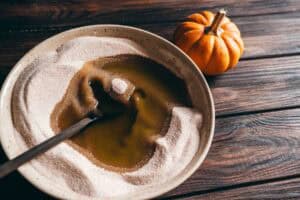 A wooden spoon stirs melted butter into a bowl of sugar next to a small pumpkin on a wooden table.