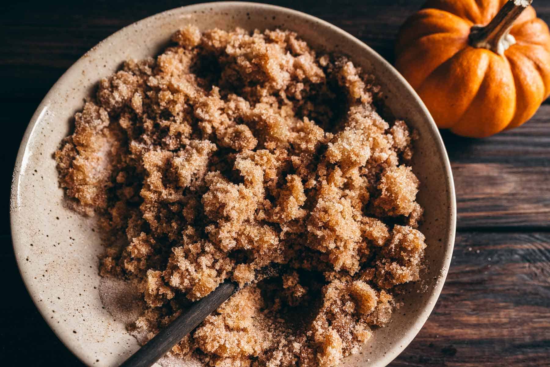 A bowl of pumpkin spice sugar scrub next to a small pumpkin on a wooden surface.