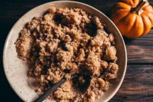 A bowl of brown sugar mixture with a wooden spoon, next to a small pumpkin on a wooden surface.