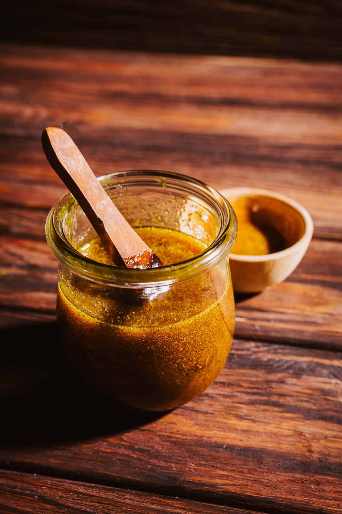 A glass jar of yellow-gold fire cider electuary with a wooden spoon, sitting on a wooden surface next to a small wooden bowl.