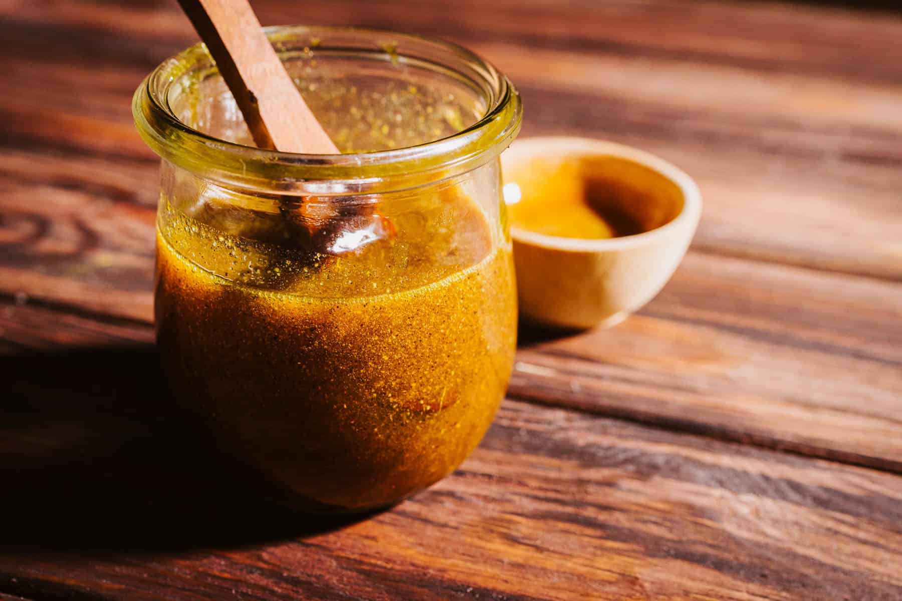 A glass jar of yellow-gold fire cider electuary with a wooden spoon, next to a small wooden bowl on a wooden surface.