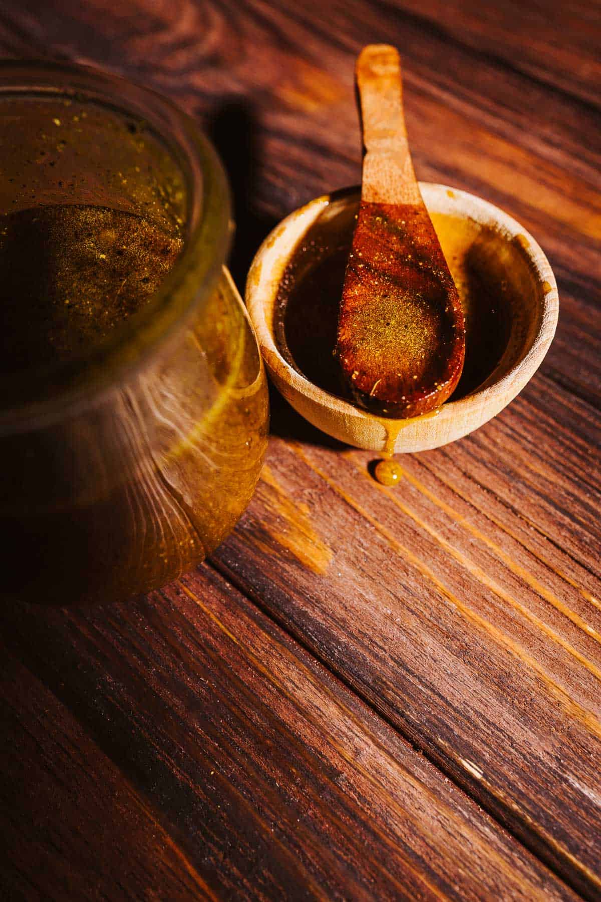 A glass jar of dark fire cider electuary next to a small wooden bowl and spoon containing the same liquid.