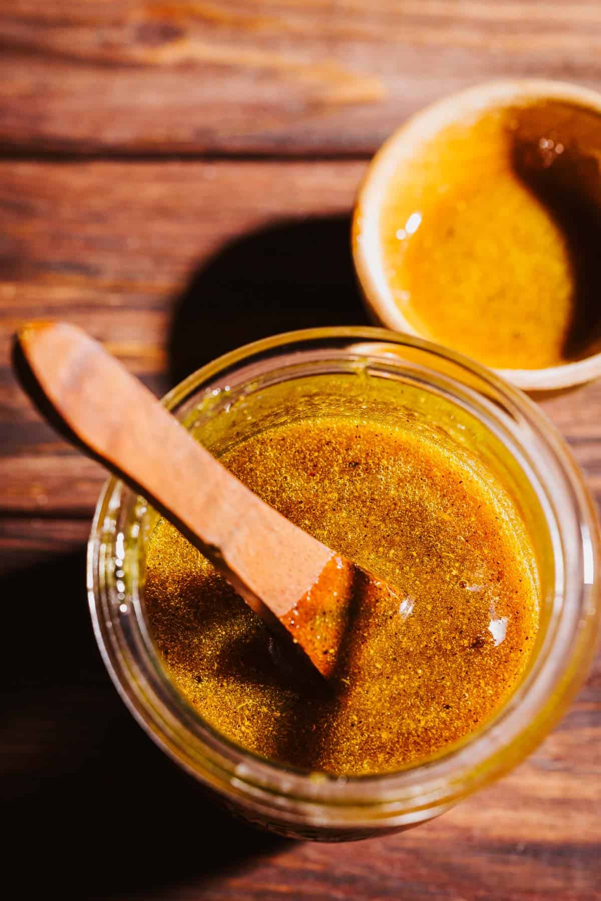 A wooden spoon rests in a glass jar of golden, spiced fire cider electuary, with a small bowl of the same mixture nearby.
