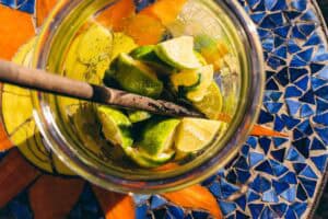 A glass jar filled with lime wedges and mint leaves, with a wooden muddler resting inside, set on a blue and orange mosaic table.
