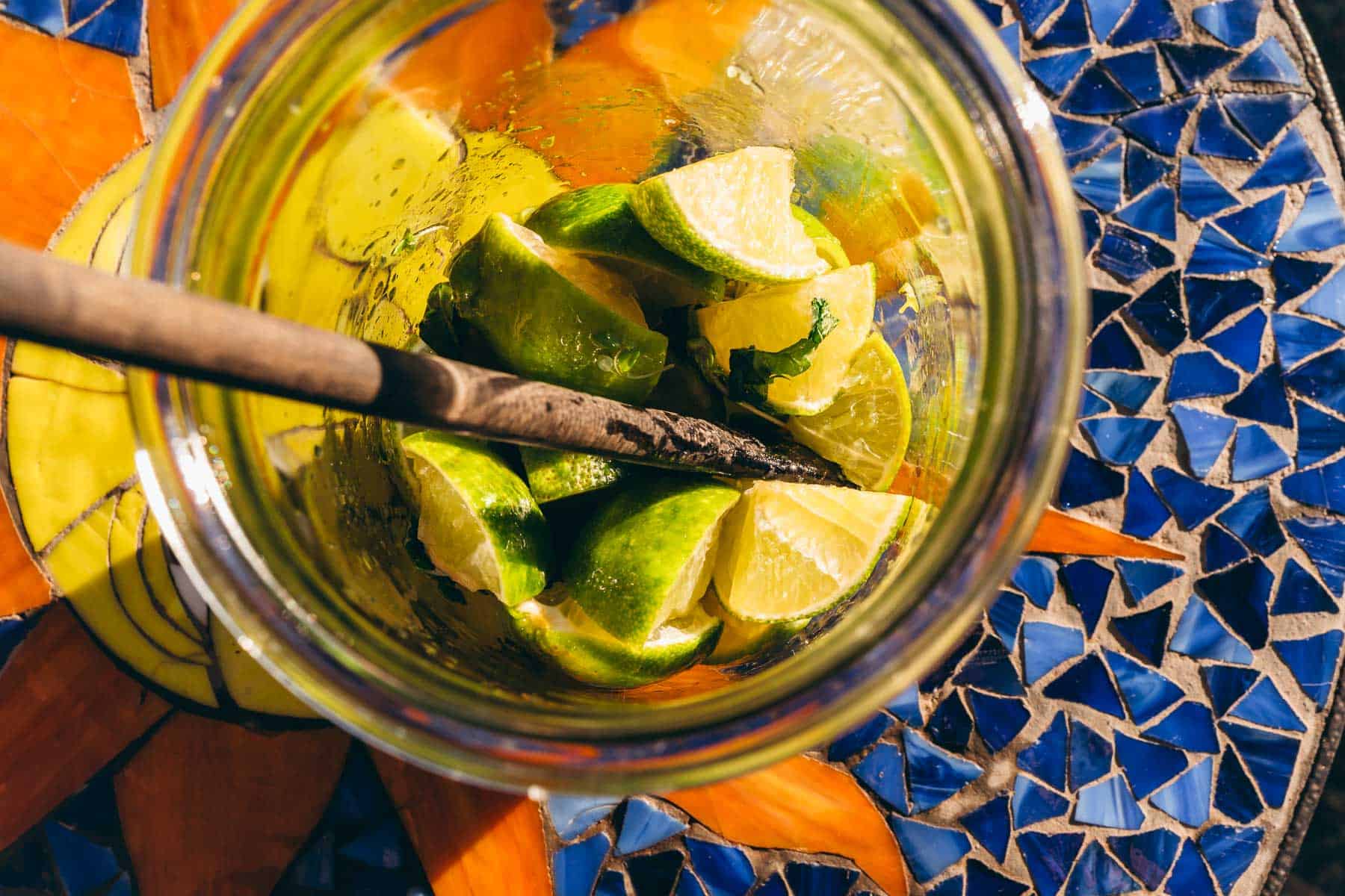 A glass jar filled with lime wedges and mint leaves, with a wooden muddler resting inside, set on a blue and orange mosaic table.