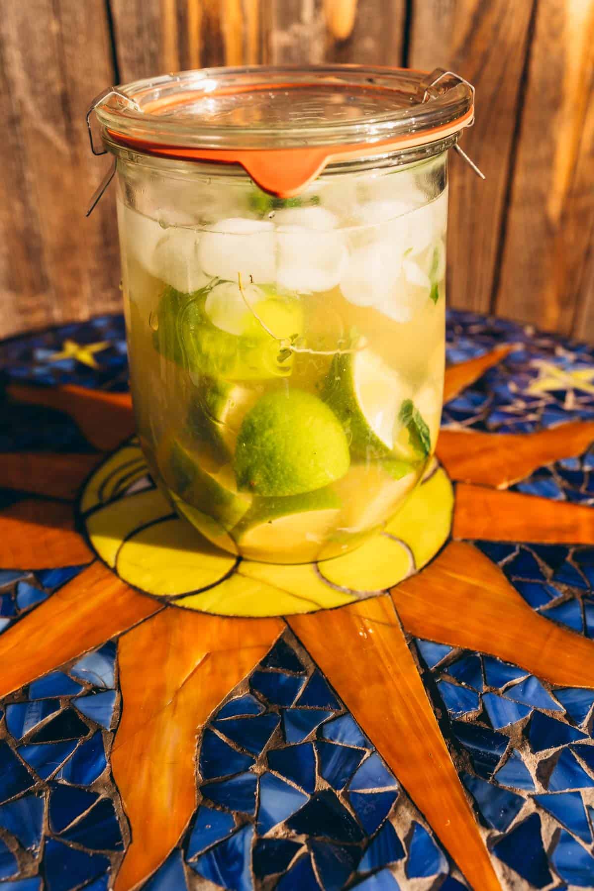 A glass jar filled with lime wedges, ice, and liquid sits on a mosaic table.