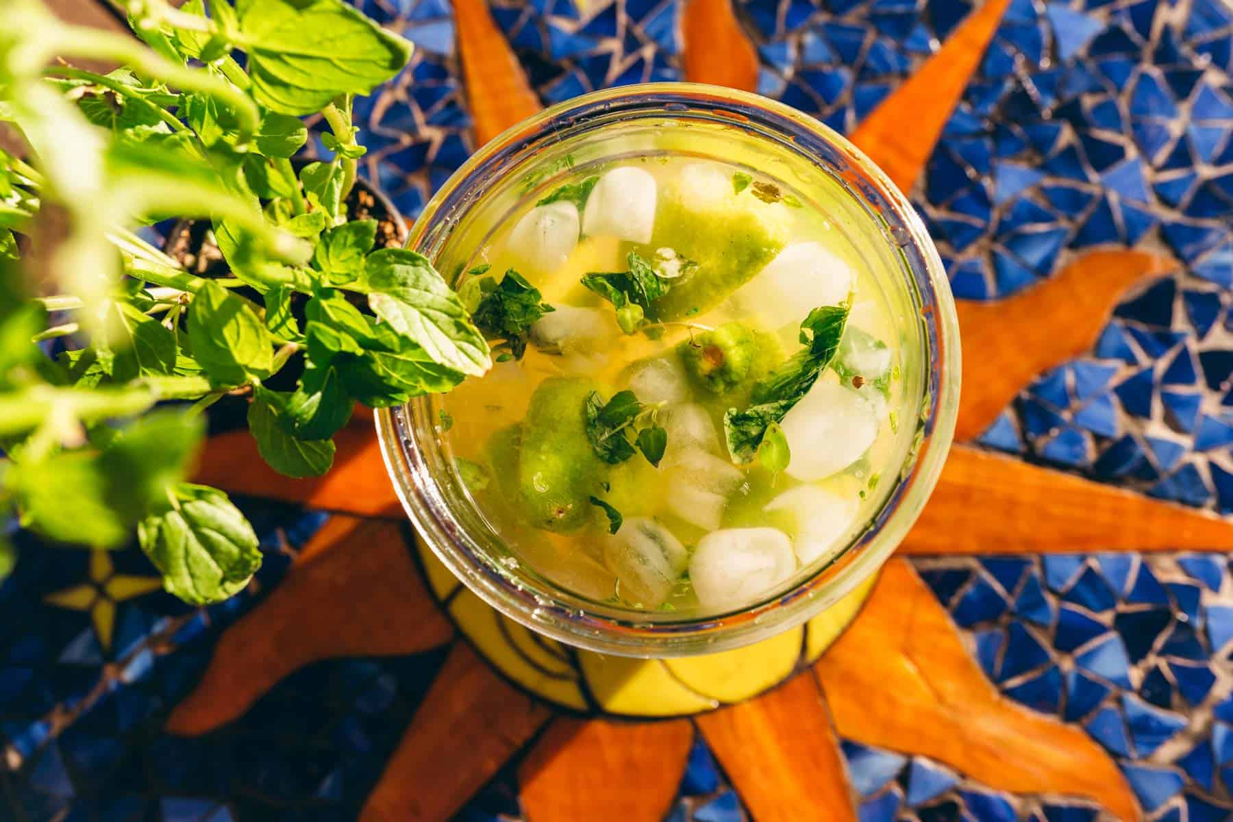 Top-down view of a glass of herbal limeade with mint and ice, placed on a tiled table with a sunburst pattern.