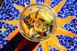 A glass jar of fire cider ingredients, including lemon slices, orange peel, rosemary sprigs, and a cinnamon stick, sitting on a mosaic table with a sun pattern.
