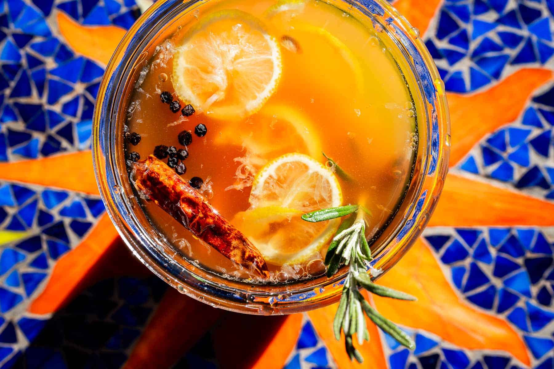 A glass of fire cider garnished with lemon slices, black peppercorns, a dried chili, and a sprig of rosemary.