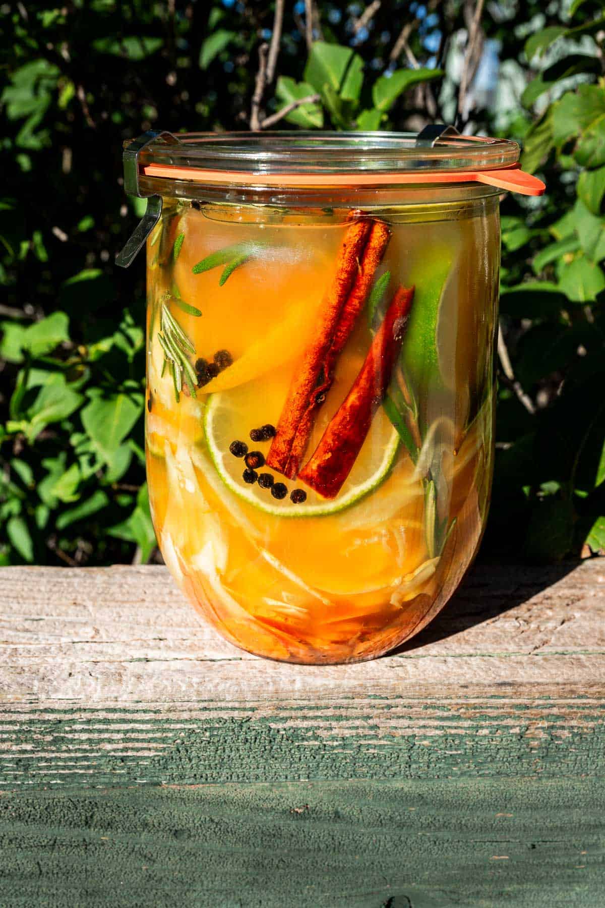 A glass jar of fire cider filled with a yellow liquid, lime slices, cinnamon sticks, peppercorns, and rosemary sprigs.