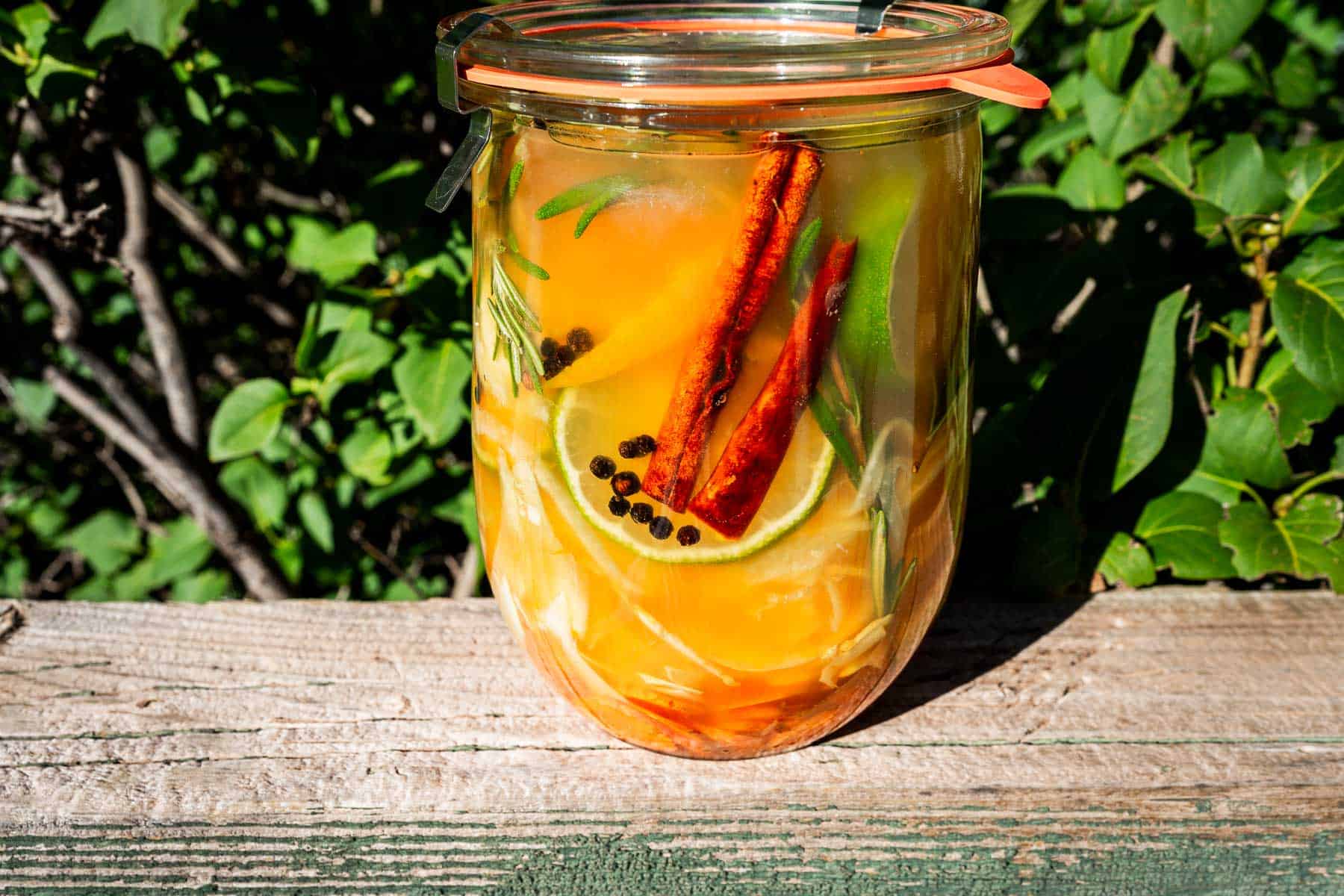 A glass canning jar filled with fire cider ingredients, including liquid, citrus slices, cinnamon sticks, peppercorns, and sprigs of rosemary.