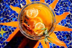 Overhead view of a glass of amber-colored fire cider garnished with lemon slices, peppercorns, a cinnamon stick, and a sprig of rosemary.