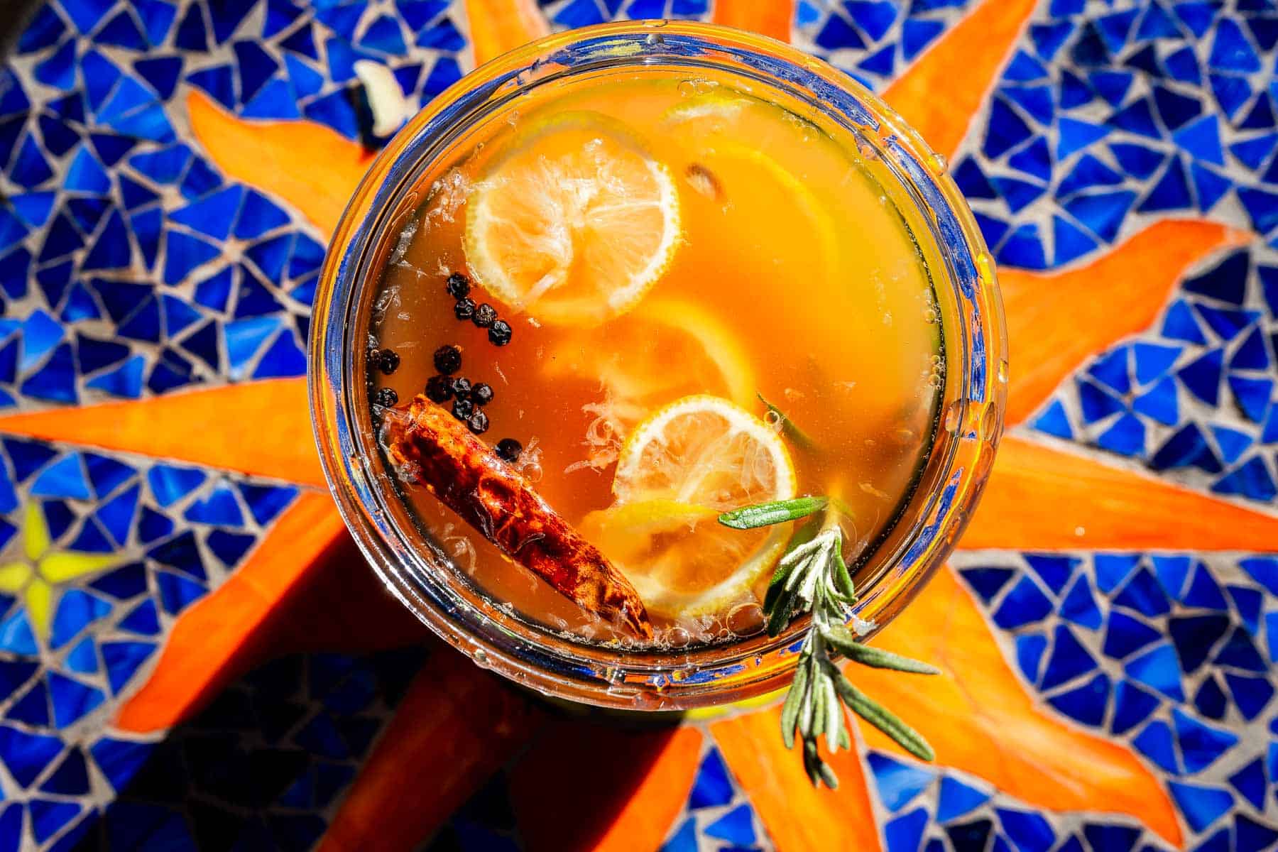 Overhead view of a glass of amber-colored fire cider garnished with lemon slices, peppercorns, a cinnamon stick, and a sprig of rosemary.