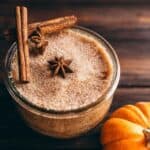 A glass jar filled with pumpkin spice sugar scrub, garnished with two cinnamon sticks and two star anise, next to a small pumpkin.